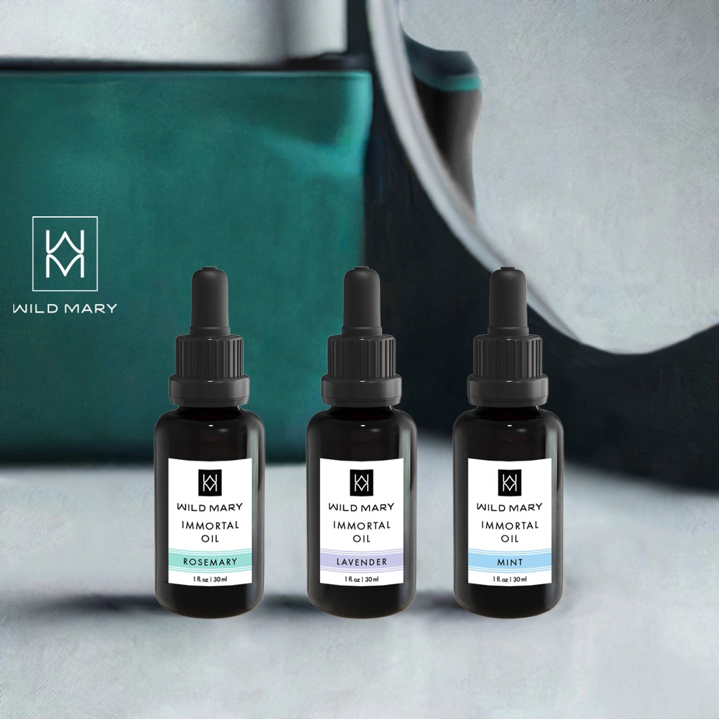 Wild Mary Immortal Oil Trio — Rosemary, Lavender, and Mint bottles displayed on a vanity with a teal bag in the background.